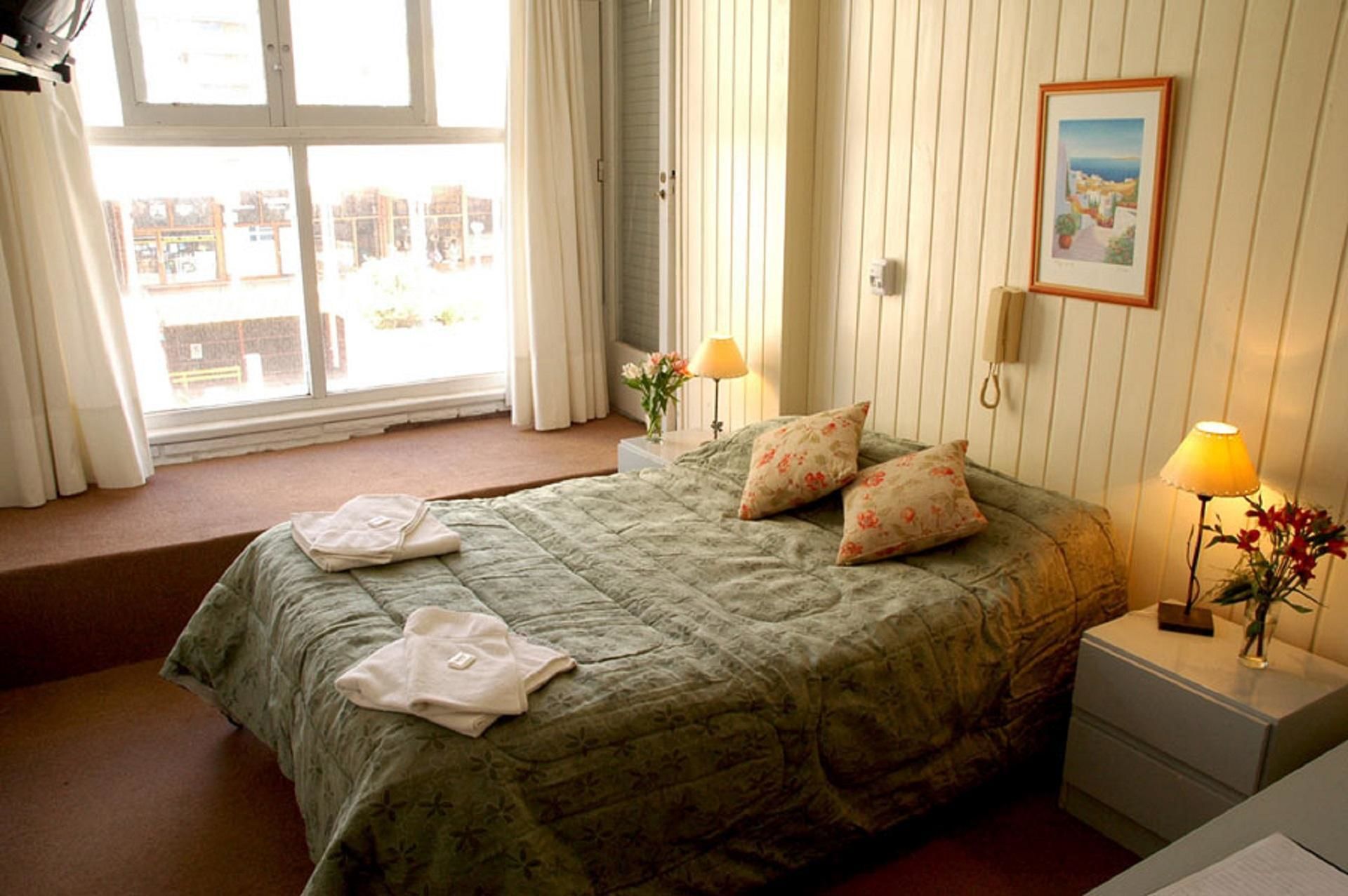 Un dormitorio con una cama, dos lámparas de noche, una mesita de noche, una ventana con cortinas y un cuadro en la pared.