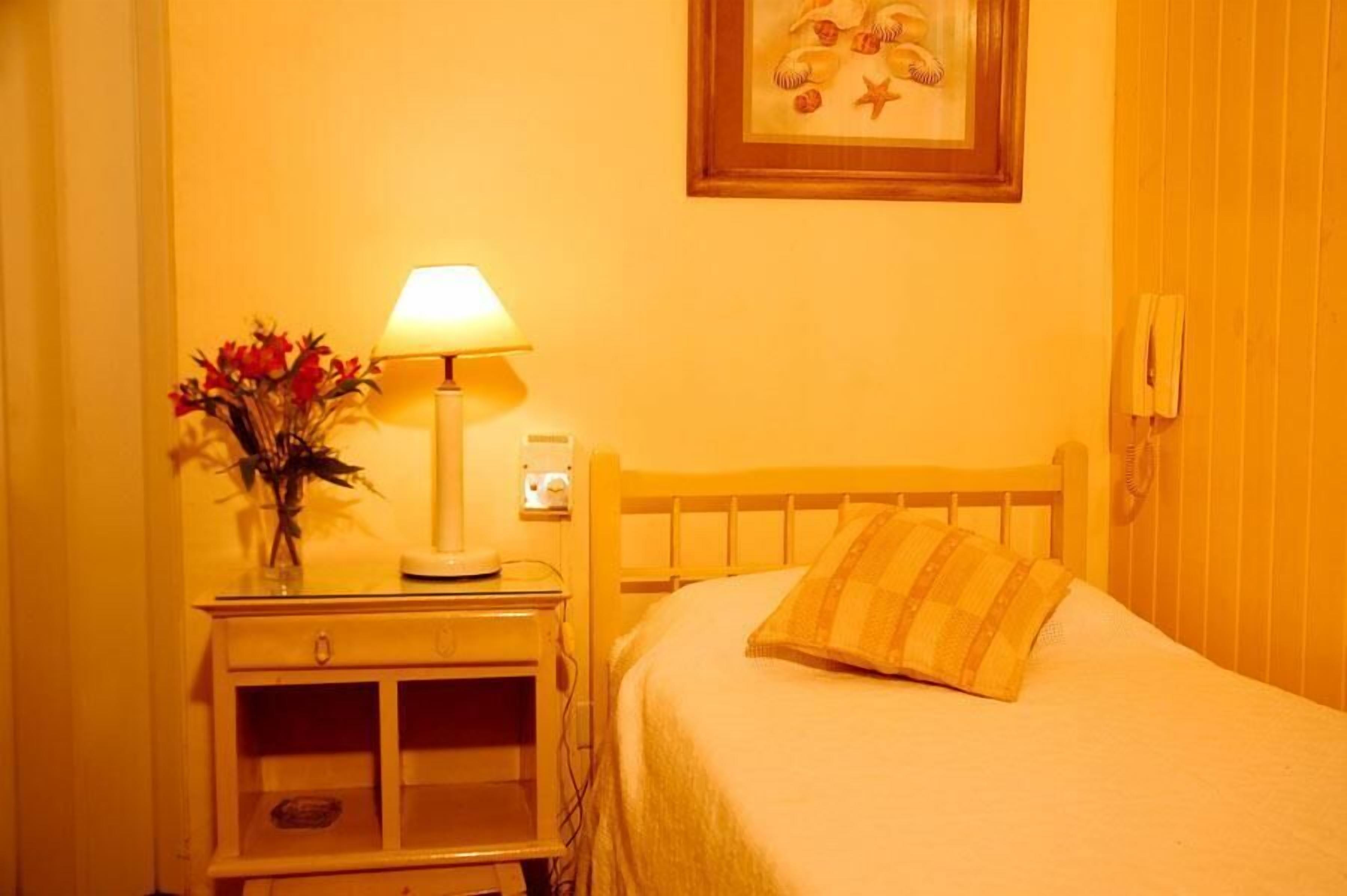 Une chambre à coucher avec un lit, une table de chevet avec une lampe et un vase de fleurs, et un tableau encadré au mur.
