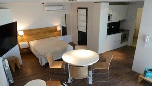 Habitació d'hotel petita i funcional, amb un llit, una taula rodona, dues cadires, una televisió, una làmpada de nit, un panell de fusta al capçal del llit, una cuineta i un terra de fusta.