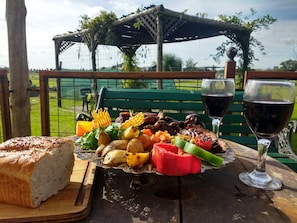 Un montaje para picnic con diversos alimentos, como pan, frutas, verduras y carne, sobre una mesa de madera con vistas a un parque y un pabellón al fondo.