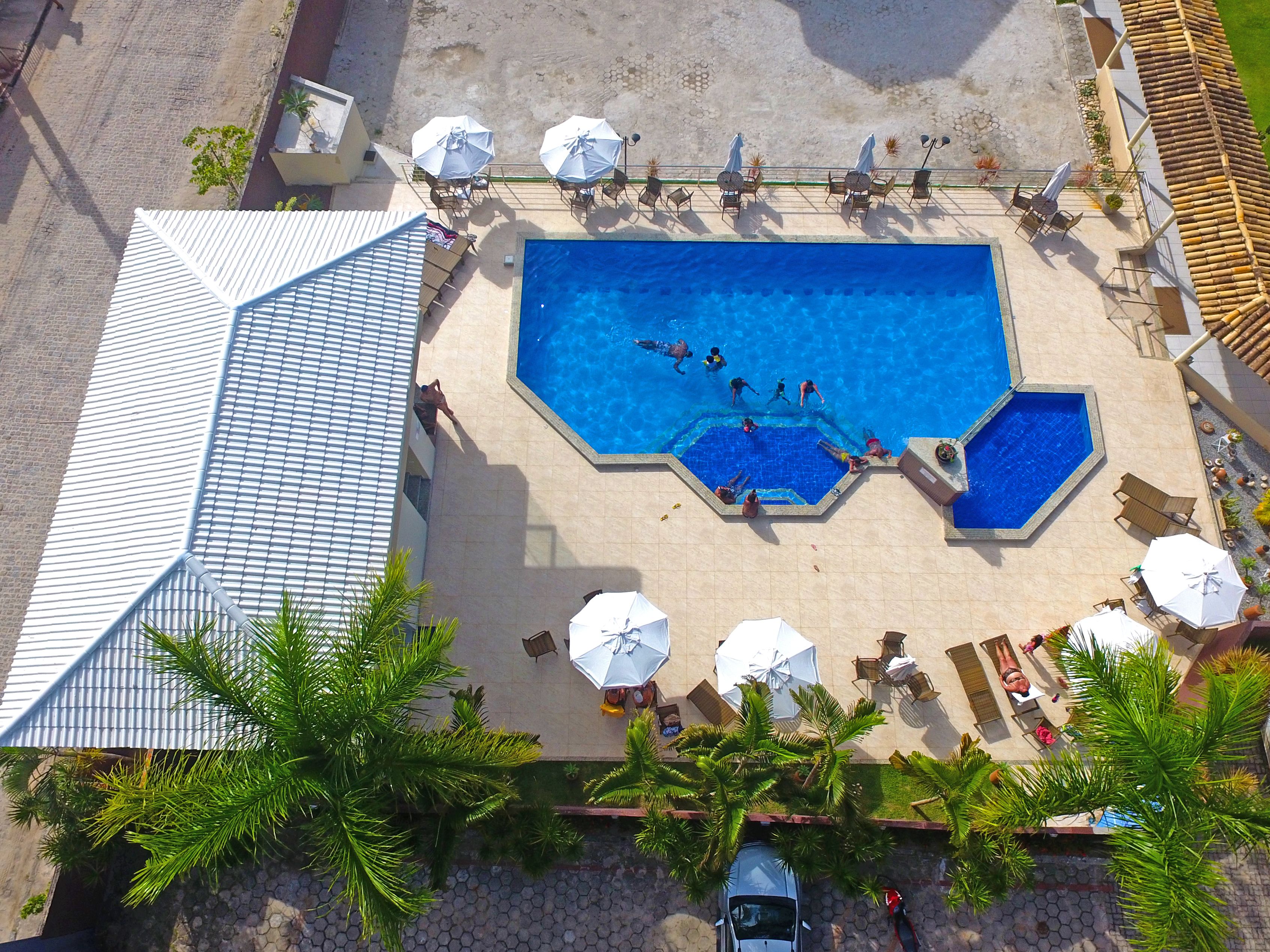 Une vue aérienne d’une piscine dotée de plusieurs couloirs, entourée de chaises longues et de parasols, et bordée de palmiers.