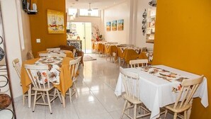 A dining area with multiple tables and chairs, featuring a bright interior with a yellow accent wall and artwork on the walls. - Residencial Ilha Dourada (Florianópolis)