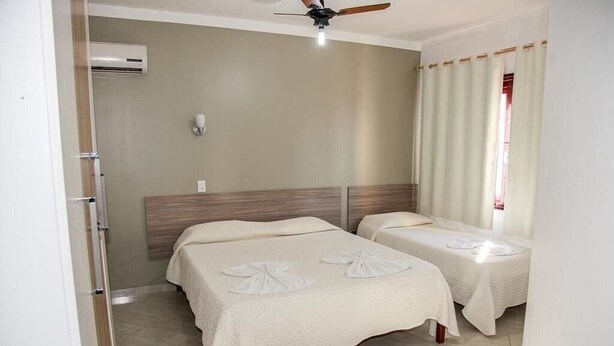 A hotel room with two beds, a ceiling fan, an air conditioning unit, and a window with curtains. - Residencial Ilha Dourada (Florianópolis)