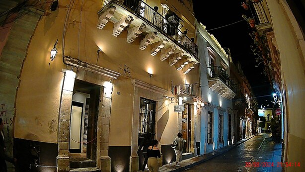 Une vue de rue de nuit, avec un immeuble doté d’un balcon et d’une enseigne sur laquelle on peut lire « ALTO ».
