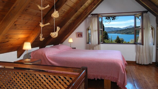Un dormitorio acogedor con techo de madera, una cama con colcha rosa, una mesita de noche con lámpara y una ventana que ofrece una vista panorámica de un lago y montañas.