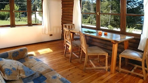 Un interior de cabaña acogedor con una mesa de comedor de madera, dos sillas, un sofá con cojines estampados y ventanas amplias que ofrecen vistas al exterior.