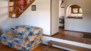 Salón con un sofá estampado, una escalera de madera y una cocina visible a través de un arco.