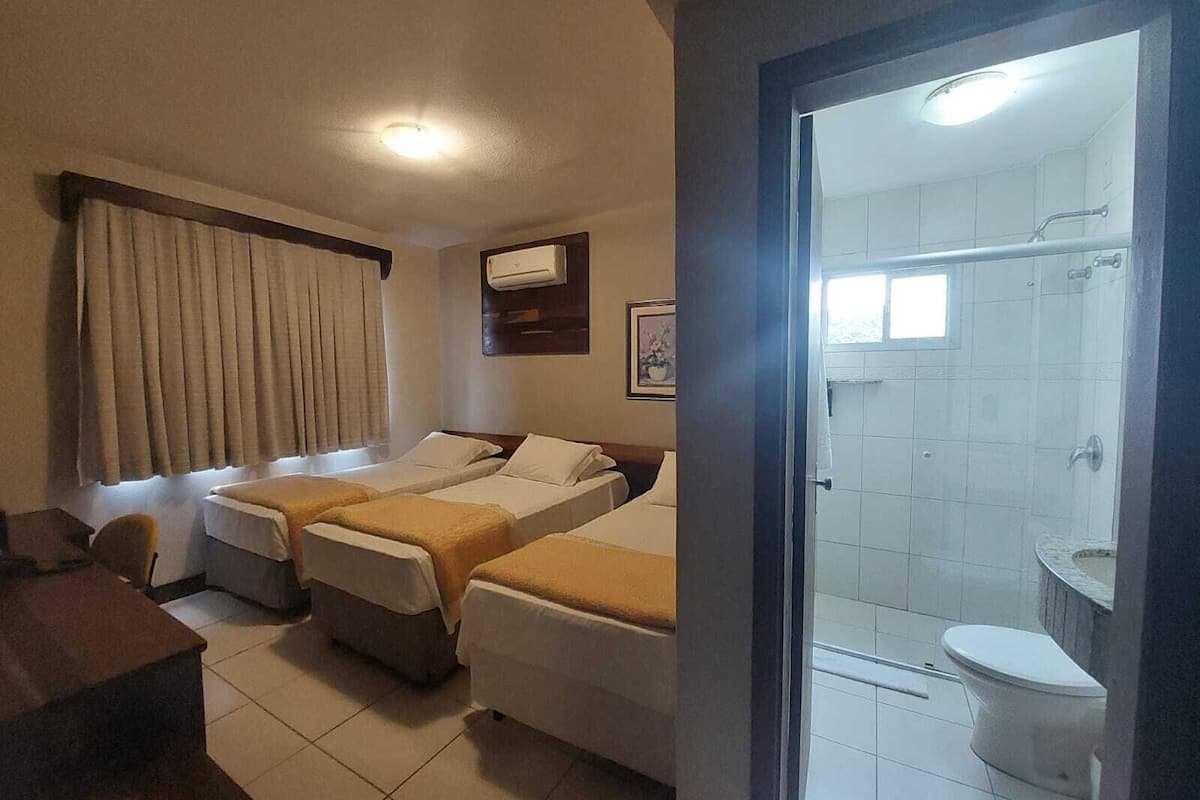 A hotel room with two beds, a desk, a window with curtains, an air conditioning unit, and a bathroom with a shower and toilet.