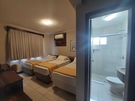 Quarto de hotel com duas camas, uma escrivaninha, janela com cortinas, ar condicionado e banheiro com chuveiro e vaso sanitário.