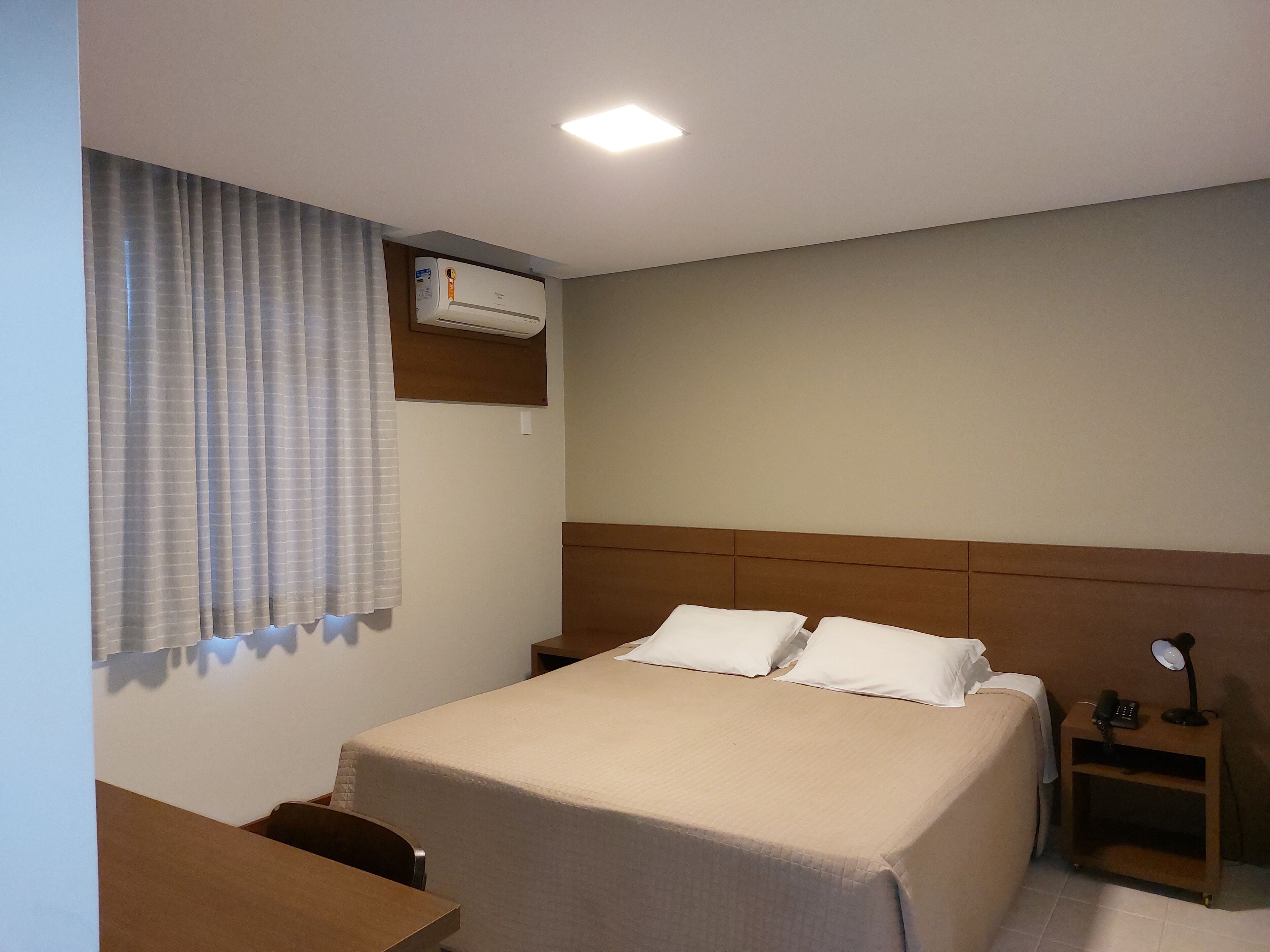 A hotel room with a bed, a desk, a chair, an air conditioning unit, a nightstand with a lamp, and a window with curtains.