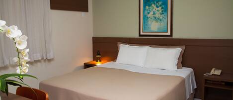 A hotel room with a bed, a nightstand, a lamp, a telephone, a painting on the wall, and a potted orchid.