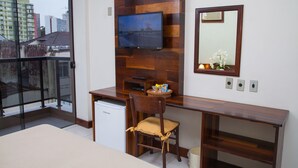A hotel room with a wooden desk, a television mounted on the wall, a chair, a small refrigerator, and a balcony with a view of the city. - Hotel Príncipe (Joinville)