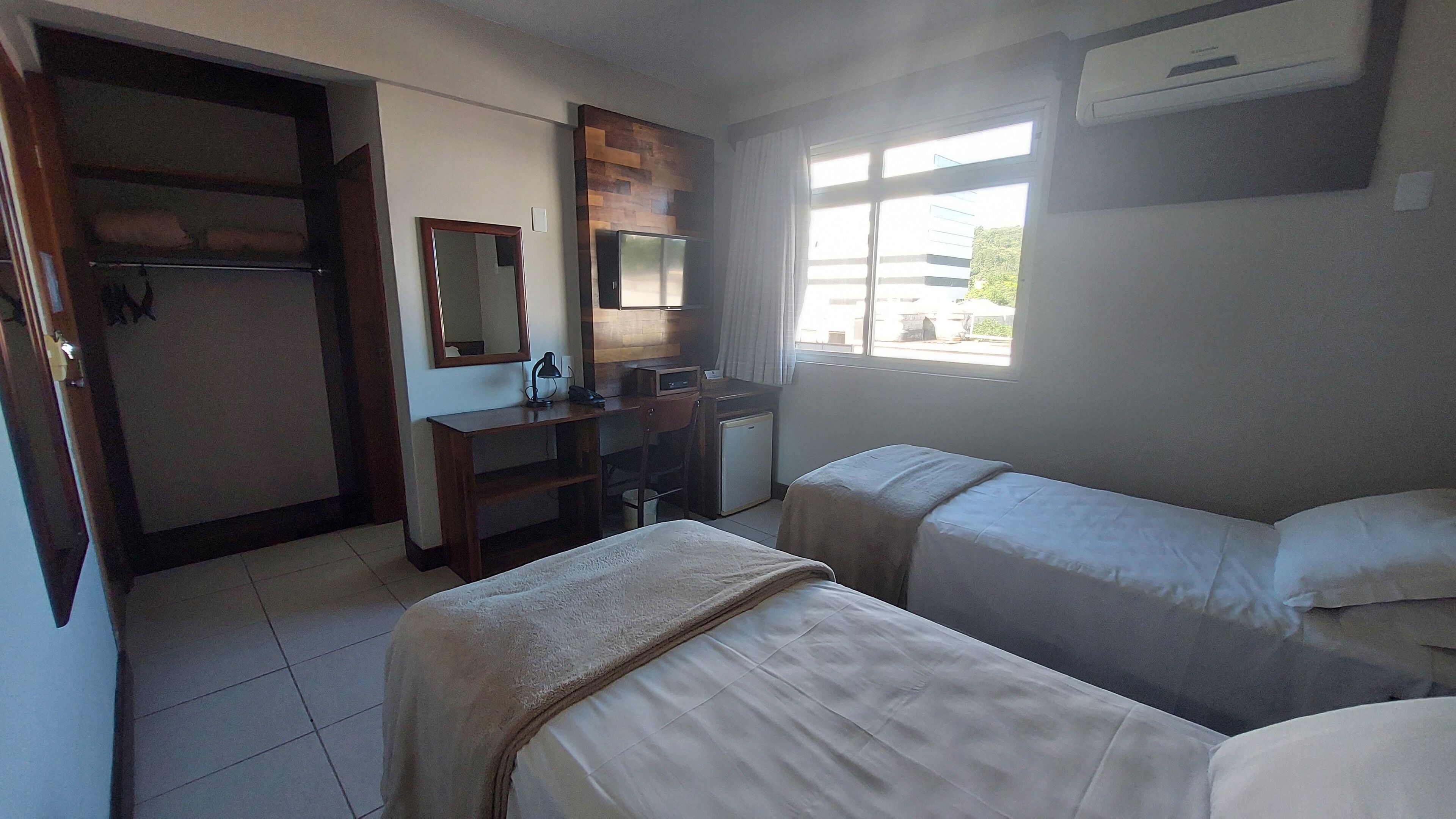 Habitación de hotel con dos camas, un escritorio con silla, un refrigerador pequeño, un espejo y una ventana con vista al exterior.