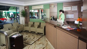 A hotel reception area with a receptionist, seating area, and a view of the street outside. - Hotel Príncipe (Joinville)