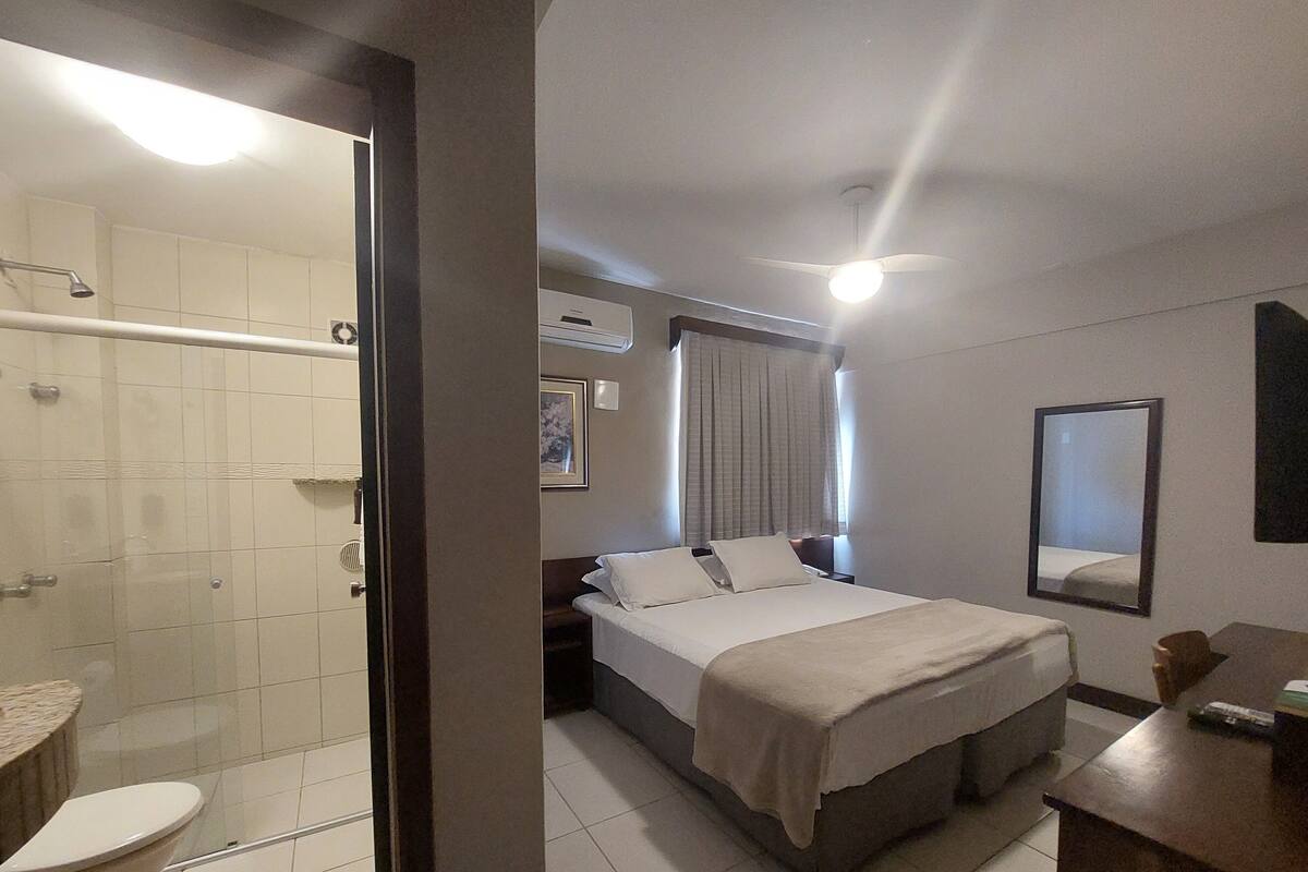 A hotel room with a bed, a bedside table, a mirror, and a shower area.