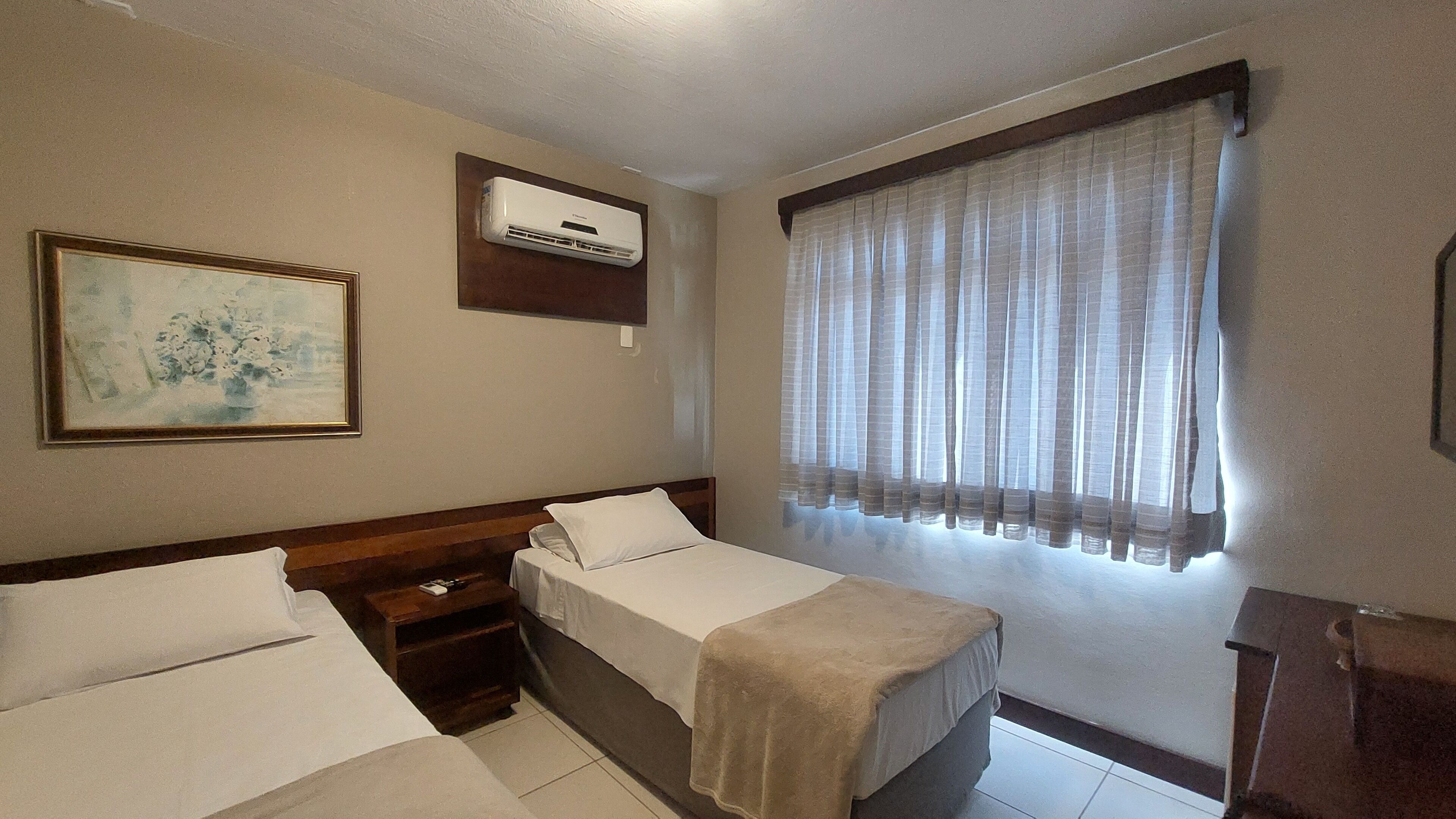 A hotel room with two single beds, a wooden headboard, a nightstand, a small table, an air conditioning unit, a window with sheer curtains, and a framed painting on the wall.