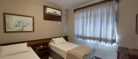 A hotel room with two single beds, a wooden headboard, a nightstand, a small table, an air conditioning unit, a window with sheer curtains, and a framed painting on the wall.