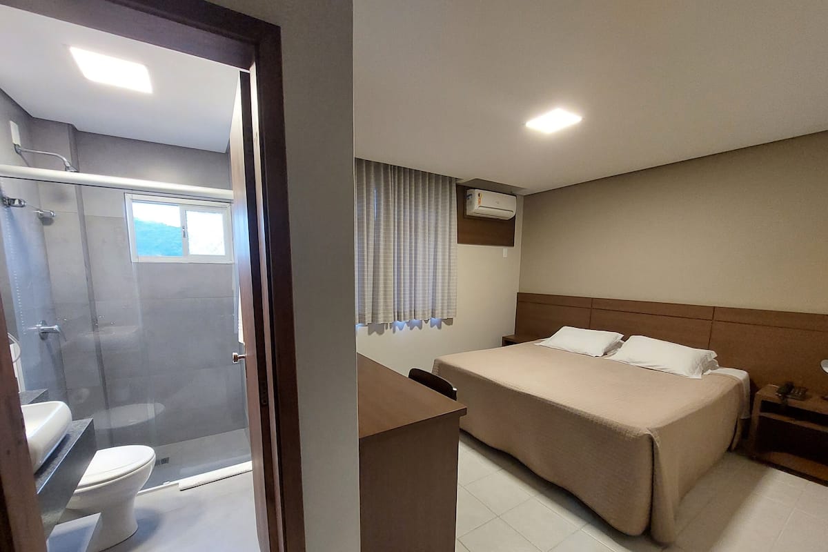 A hotel room with a shower, toilet, bed, bedside table, and a window with curtains.