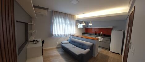 A modern kitchen with a built-in microwave, a refrigerator, and a countertop with a sink. There is a grey sofa in the living area and a window with curtains.