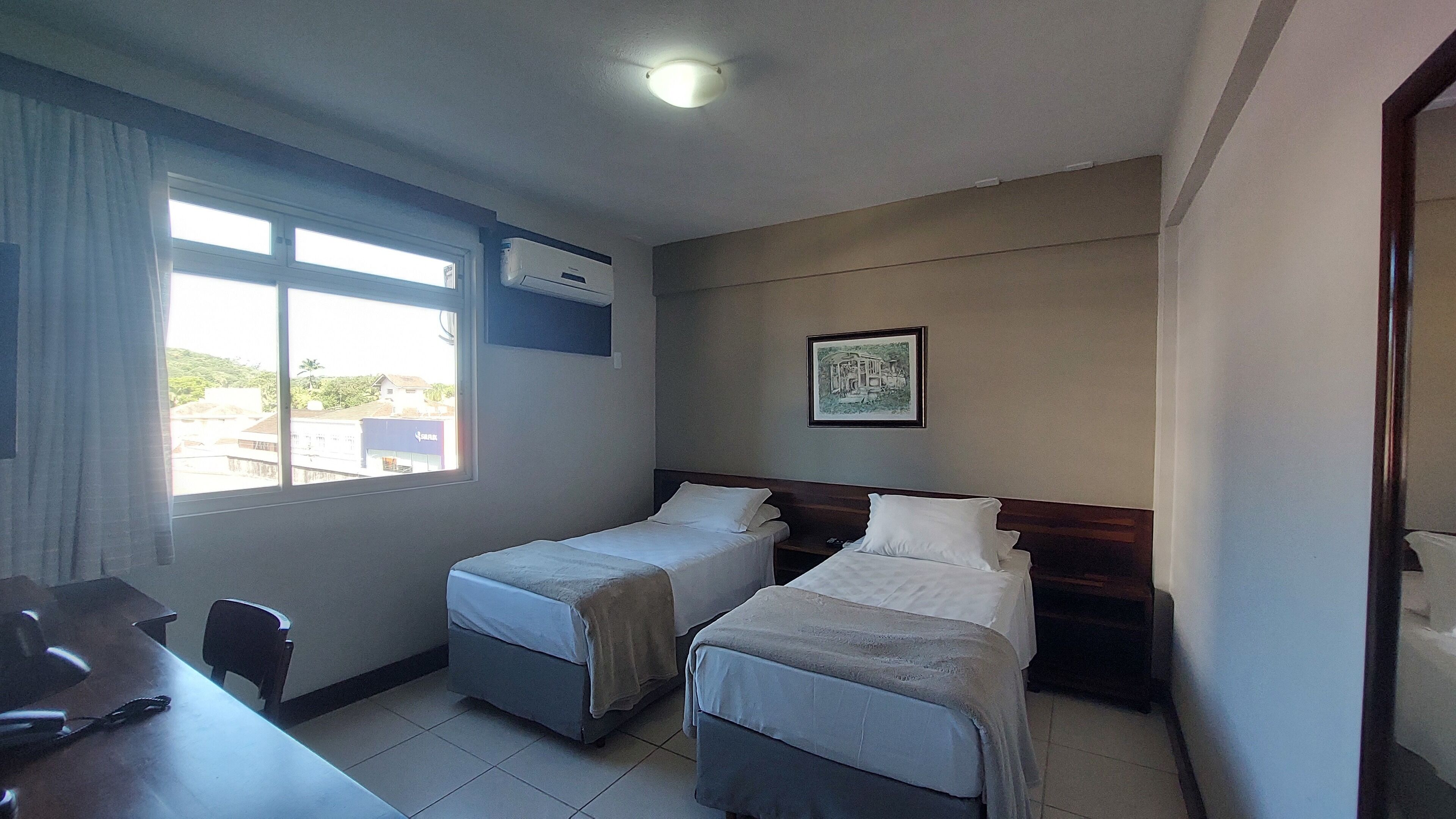 A hotel room with two single beds, a desk, a chair, a window with curtains, an air conditioning unit, and a framed picture on the wall.