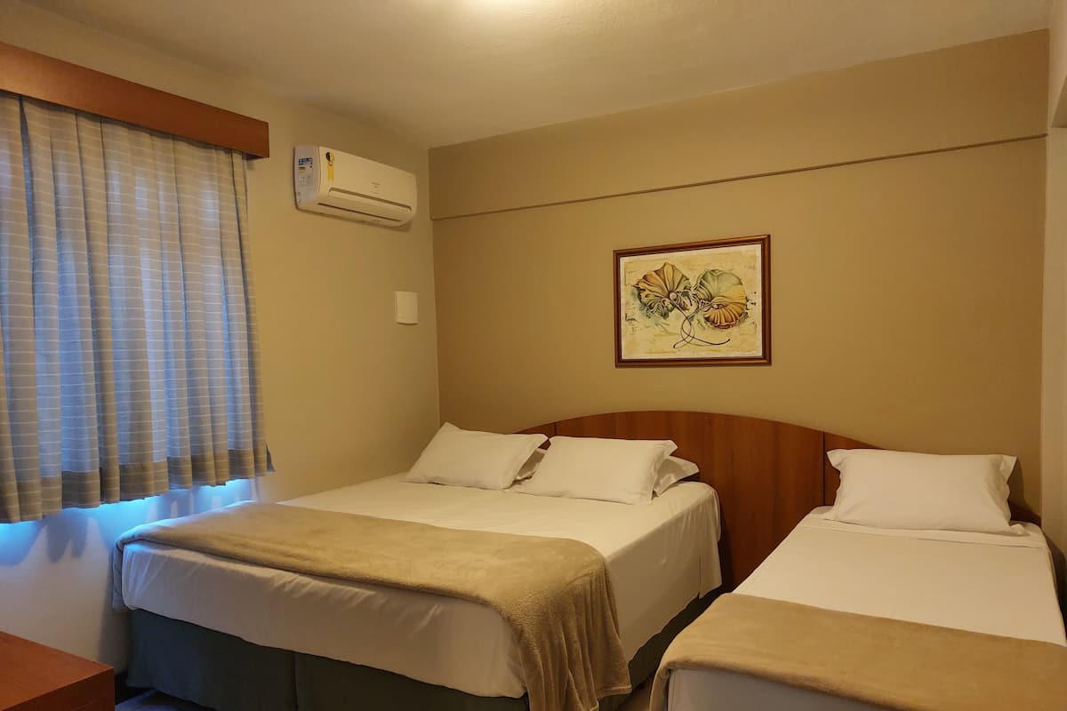 A hotel room with two beds, a window with curtains, an air conditioning unit, and a framed picture on the wall.