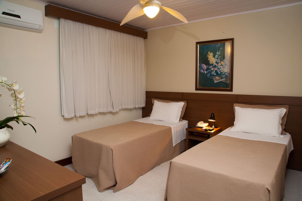 A hotel room with two single beds, a nightstand with a lamp, a painting on the wall, and an air conditioning unit.
