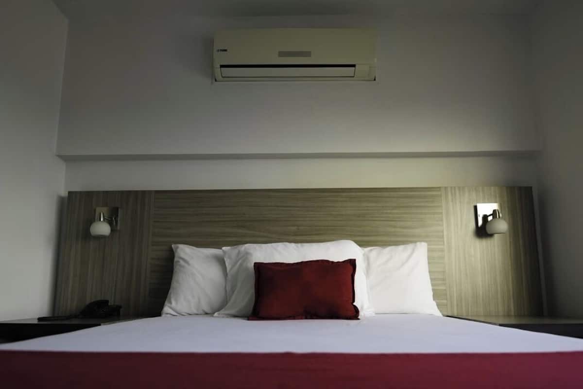 A hotel room with a bed, a red throw pillow, a white pillow, a nightstand, a lamp, and an air conditioning unit.