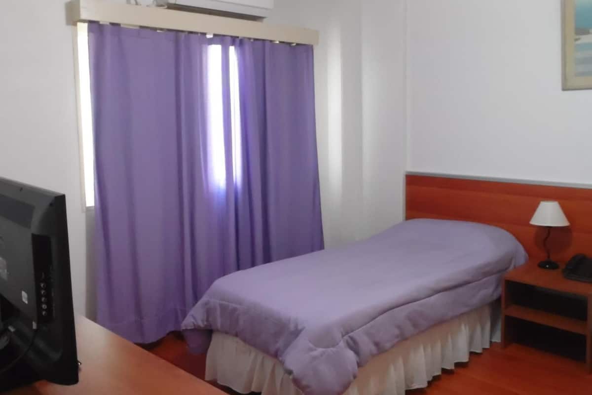 A bedroom with a bed, a desk, a chair, an air conditioning unit, and a painting on the wall.