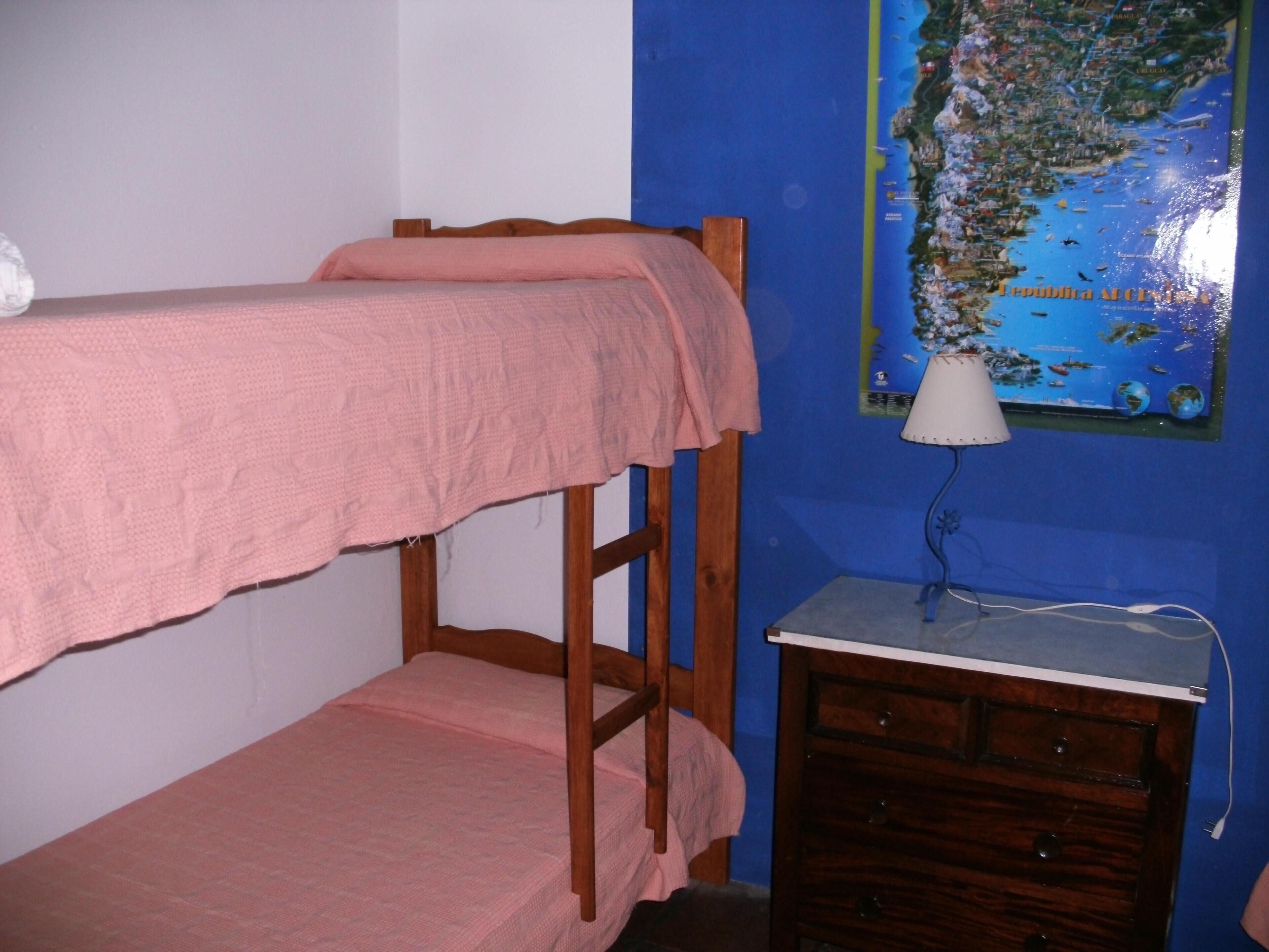 Una cama litera con una manta rosa, un pequeño aparador de madera y un mapa en la pared.