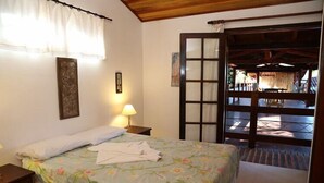 A bedroom with a floral bedspread, a nightstand with a lamp, a framed picture on the wall, and a view of an outdoor patio through a glass door. - Pousada Enseada dos Búzios (Ubatuba)
