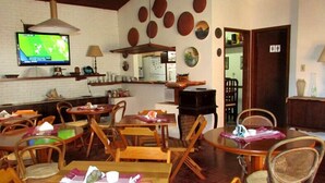 A restaurant with wooden tables and chairs, a TV displaying a sports game, and a kitchen area visible through an open door. - Pousada Enseada dos Búzios (Ubatuba)