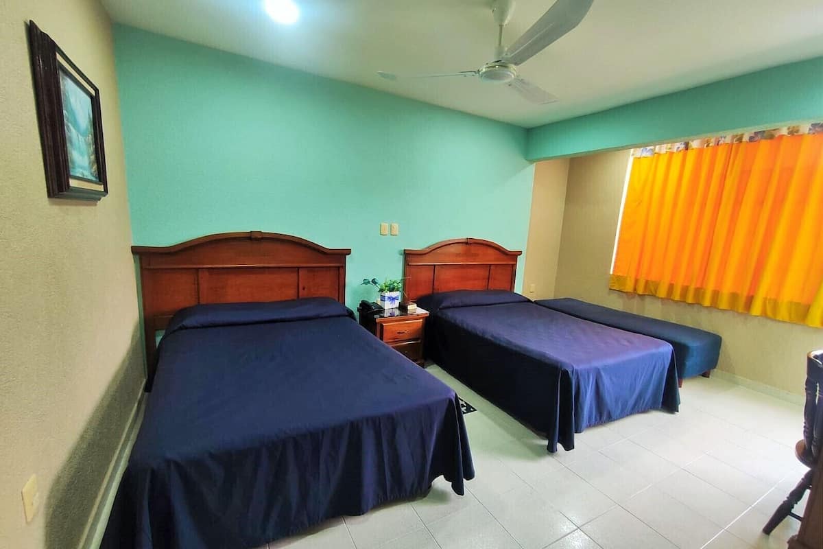 A hotel room with two single beds, a ceiling fan, a window with orange curtains, and a small table with a plant.