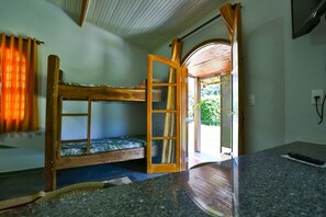 Un lit superposé avec une structure en bois et un panneau en verre, un petit coin salon avec un coussin et une télécommande posée sur un comptoir en granit.
