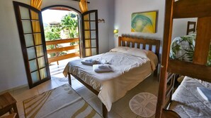 Une chambre à coucher avec un lit, une tête de lit en bois, une fenêtre avec des rideaux, un balcon et une vue sur la verdure.