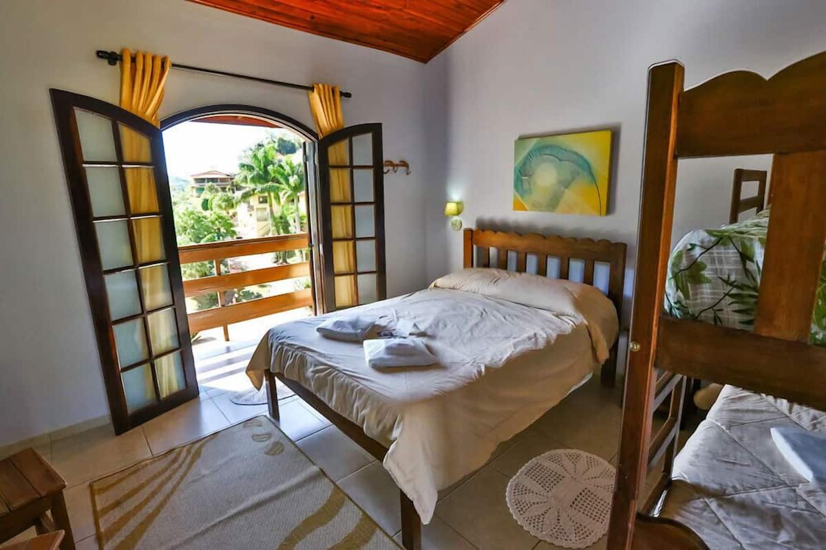 Un dormitorio con cama, cabecera de madera, ventana con cortinas, balcón y vistas al verde.