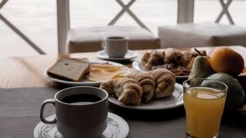 A breakfast setting with a cup of coffee, croissants, fruit, and a glass of juice on a table by a window with a view.