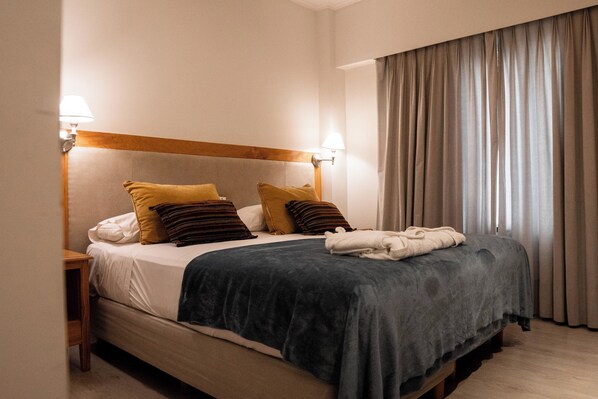 Una cama bien tendida con un edredón azul oscuro, dos almohadas y una toalla doblada. Hay dos lámparas de noche a cada lado de la cama.
