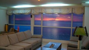 A living room with a sofa, a coffee table, and a lamp. There is a large window with a view of the sea. - Apartamentos Suiteline Master (Santa Marta)