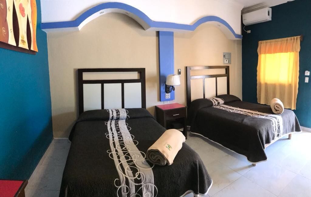 Habitación de hotel con dos camas, una mesita de noche, una lámpara y una ventana con cortinas.