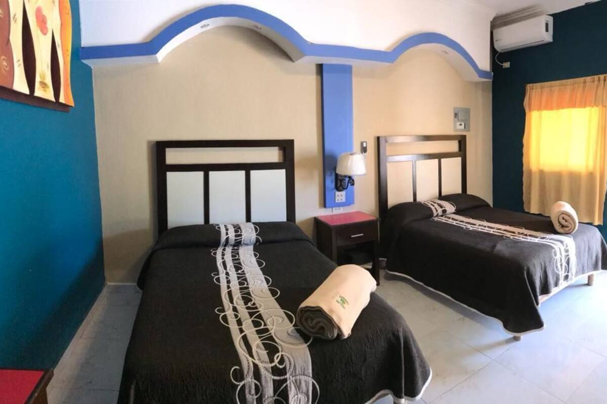 Habitación de hotel con dos camas, una mesita de noche, una lámpara y una ventana con cortinas.