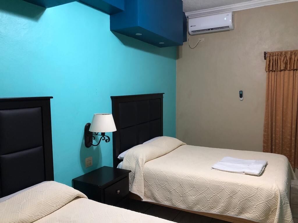 Habitación de hotel con dos camas, una mesita de noche, una lámpara de pared, un equipo de aire acondicionado y una ventana con cortinas.