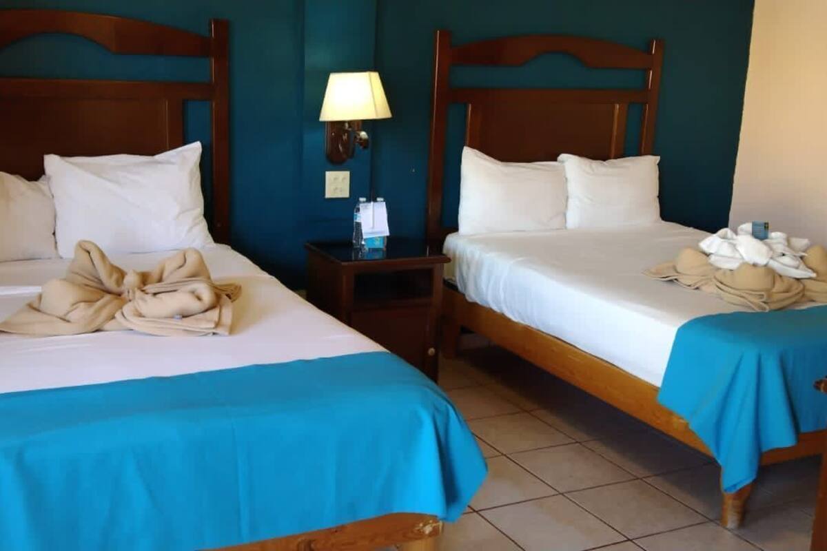 Dos camas matrimoniales con cubrecamas azules, almohadas blancas y toallas dobladas sobre cada una. Hay una mesita de noche con una lámpara entre las camas.