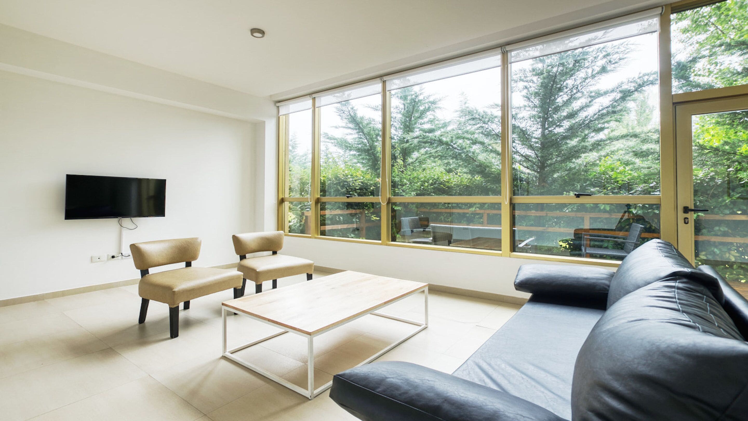 A modern living room with a large window, a flat-screen TV, two chairs, a coffee table, and a sofa.