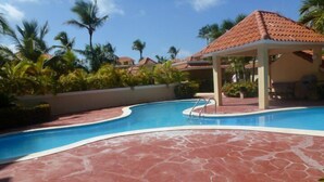 Una piscina con plataforma de baldosas rojas, rodeada de palmeras y un pabellón.