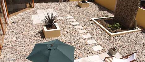 Un patio exterior con suelo de baldosas, un paraguas verde y varias sillas y macetas.