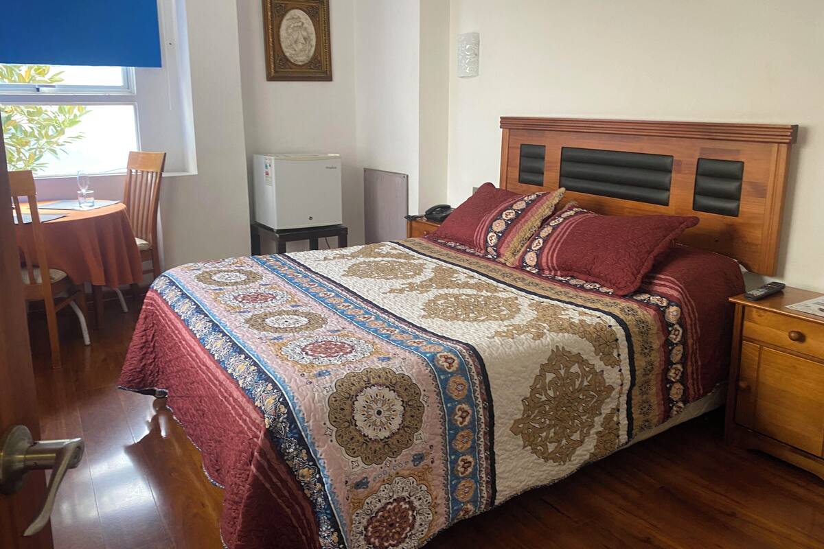 Ein Schlafzimmer mit einem hölzernen Bett, einer gemusterten Bettdecke, einem kleinen Tisch mit Stuhl, einem Kühlschrank und einem Fenster mit Jalousien.
