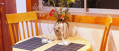 Una mesa de comedor con mantel amarillo, dos individuales cuadrados, un florero con flores y un vaso de agua.