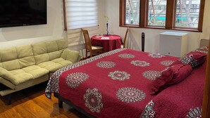 A bedroom with a bed, a sofa, a TV, a table with a red tablecloth, and a window with blinds. - Nubes Hotel (Vina del Mar)