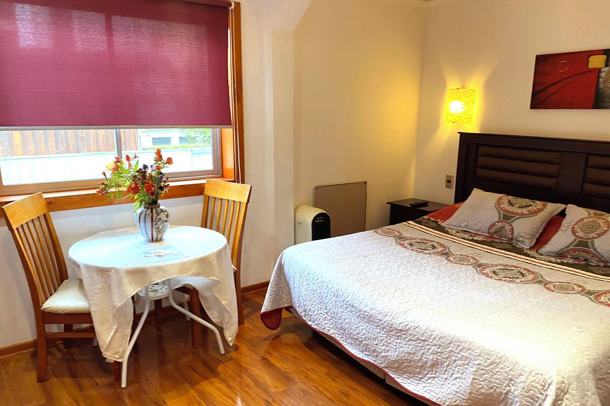 Ein Schlafzimmer mit Bett, Nachttisch, Sessel, Tisch mit Blumenvase und Fenster mit Außenblick.