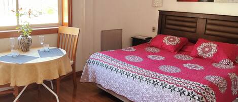 Un dormitorio con cama, una mesa y sillas, un florero con flores y una ventana con vistas.